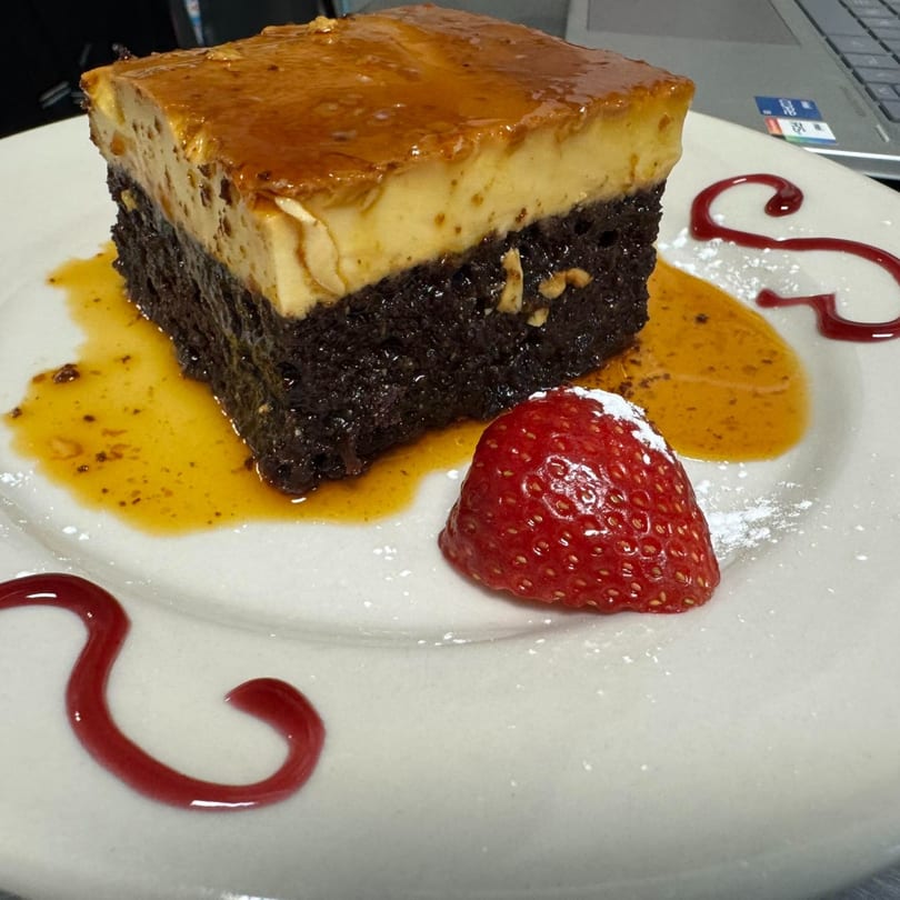 A slice of chocolate cake with flan on top, drizzled with caramel, strawberry, and raspberry sauce.